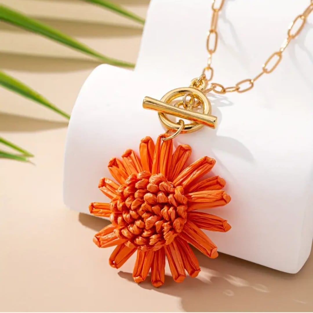 This playful piece features a vibrant orange raffia daisy as its centerpiece, radiating warmth and cheer. The flower’s textured petals bring a handcrafted, organic charm, while the delicate gold chain adds a touch of elegance and sophistication. The contrast between the rustic raffia and the refined gold creates a balanced design that feels both whimsical and chic.